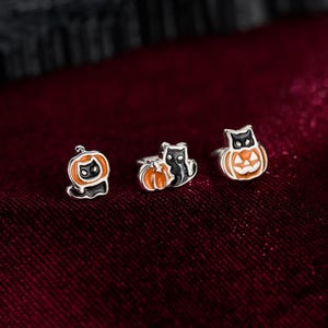 May include: Three silver-toned stud earrings featuring enamel designs. One earring shows a black cat inside an orange pumpkin, another depicts a black cat sitting next to an orange pumpkin, and the third is a black cat wearing an orange pumpkin hat.