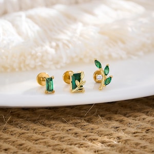 Puede incluir: Tres pendientes de oro con piedras verdes talladas en esmeralda. Uno tiene una esmeralda rectangular, otro un diseño de hoja y el tercero un diseño de hoja con una pequeña piedra blanca. Los pendientes se muestran sobre una superficie blanca.
