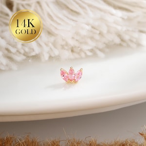 May include: A small, gold-colored earring with a floral design, featuring three pink gemstones. The earring is set on a white surface, with a gold circle that reads "14K GOLD" in the background.