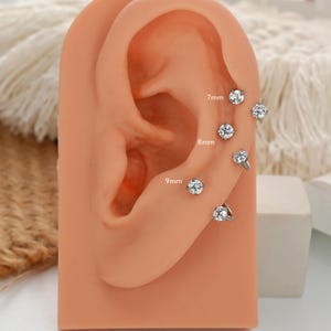 Può includere: Esposizione di diversi orecchini a bottone argentati con gemme rotonde e trasparenti. Le misure sono indicate come 7 mm, 8 mm e 9 mm. Gli orecchini sono presentati su un modello di orecchio color carne, in stile classico e semplice.