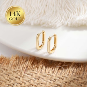 Puede incluir: Un par de pendientes de aro rectangulares dorados. Los pendientes se muestran sobre un plato blanco, con una superficie texturizada y marrón en primer plano. Un círculo dorado con el texto "14K GOLD" está en la esquina superior izquierda.