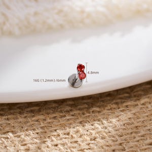 Pu&ograve; includere: Un piercing labret color argento con due piccole pietre rosse rotonde incastonate verticalmente. Il gioiello presenta una base circolare piatta e misura 16G (1,2 mm) di spessore, con una lunghezza del perno di 6 mm e 4,8 mm di altezza totale.