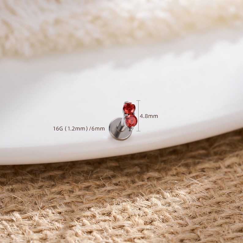Pu&ograve; includere: Un piercing labret color argento con due piccole pietre rosse rotonde incastonate verticalmente. Il gioiello presenta una base circolare piatta e misura 16G (1,2 mm) di spessore, con una lunghezza del perno di 6 mm e 4,8 mm di altezza totale.