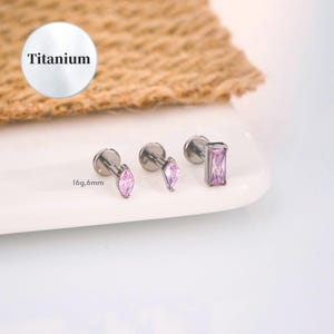 Puede incluir: Tres piercings de labio de titanio con piedras preciosas de color púrpura claro. Un pendiente tiene una gema en forma de marquesa, otro tiene una gema en forma de diamante y el tercero tiene una gema rectangular. El texto "Titanium" está en un círculo blanco.