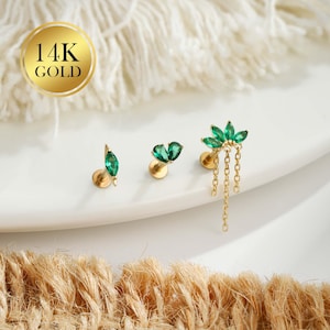 Puede incluir: Tres pendientes de oro de 14K con detalles de piedras preciosas verdes. Los pendientes presentan varios diseños, incluyendo una hoja, un corazón y un grupo floral con cadenas colgantes. Se exhiben sobre una superficie blanca con un sello dorado que dice "14K GOLD".