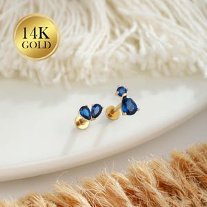 May include: Two pairs of 14K gold earrings with blue teardrop-shaped gemstones. One pair has three gemstones, the other two. The earrings are displayed on a white surface with a gold circle that says "14K GOLD".