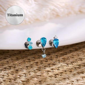 Könnte beinhalten: Drei Schmuckstücke aus Titan mit blauen Edelsteinen. Eines hat drei kleine runde Edelsteine, ein anderes einen tropfenförmigen Edelstein mit einem hängenden Edelstein, und das dritte einen tropfenförmigen Edelstein mit einem kleinen weißen Edelstein. Das Wort "Titanium" steht in einem weißen Kreis.