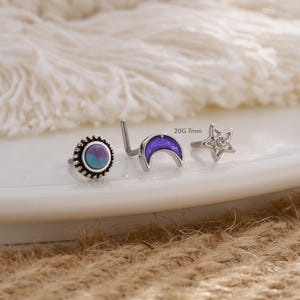 May include: Three nose rings with different designs: a round ring with a blue and purple stone, a crescent moon ring, and a star ring with a clear stone. The rings are 20 gauge and 7 mm in size.