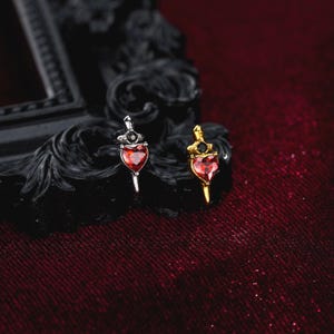 May include: Two dagger-shaped earrings, one in silver tone and one in gold tone, each featuring a faceted heart-shaped red gemstone at the center and a small black stone set above the heart. The metal daggers have pointed tips and detailed hilts.