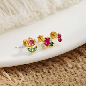 May include: Three gold-colored stud earrings with colorful gemstones. One earring features a flower design with a pink center and green leaves. Another has a star shape with a pink teardrop gem, a white gem, and a green gem. The third has two pink gems.