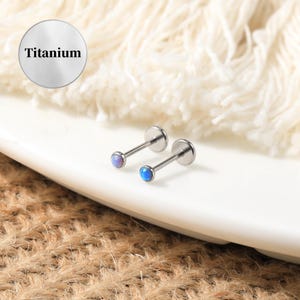 May include: Two titanium body jewelry pieces with opal stones. One stone is a light blue color and the other is a light purple color.