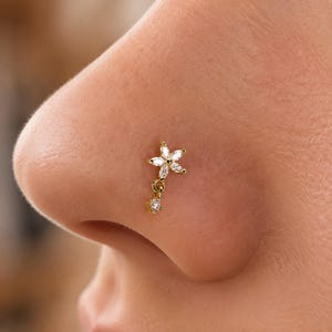 May include: A gold-toned nose stud featuring a flower-shaped design with five marquise-cut clear stones. A small jump ring connects the flower to a single dangling round clear stone. The jewellery is positioned on the side of a human nose.