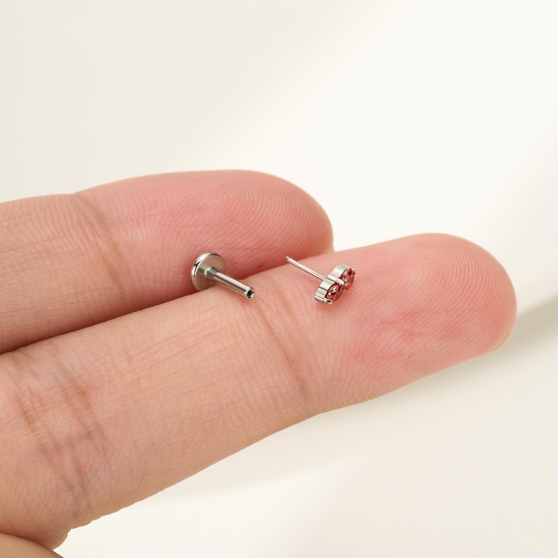 Pu&ograve; includere: Un piercing labret color argento con retro piatto, caratterizzato da una parte superiore decorativa con due piccole pietre preziose rosse rotonde incastonate in una montatura metallica. Il gioiello ha un perno dritto e una base circolare.