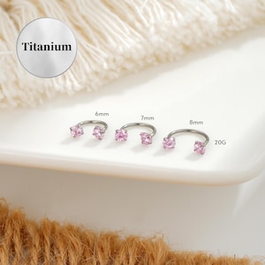 Puede incluir: Tres pendientes de herradura de titanio con piedras rosas. Las tallas son 6 mm, 7 mm y 8 mm, con un calibre de 20G. La palabra "Titanium" está en un círculo plateado.