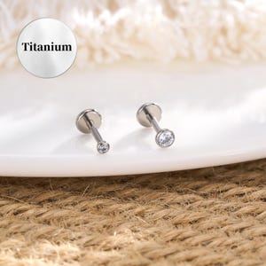 May include: Two silver-colored labret studs with clear, round gemstones. The studs are displayed on a white surface, with a circular label that reads "Titanium". The background is a neutral, textured surface.