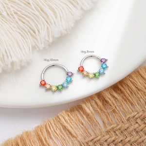 May include: Two silver-colored nose rings with a rainbow of colorful gemstones. One ring is labeled "16g,10mm" and the other is labeled "16g,8mm". The gemstones are arranged in a semi-circle around the ring.