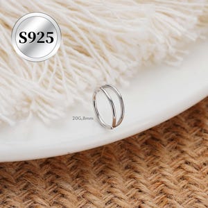 以下が含まれることがあります： シルバーのダブルフープノーズリング。リングは光沢のある金属製で、表面は滑らかです。画像には「S925」の文字が入った丸いタグが含まれています。リングのゲージは20G、直径8mmです。リングは白い表面に表示されています。