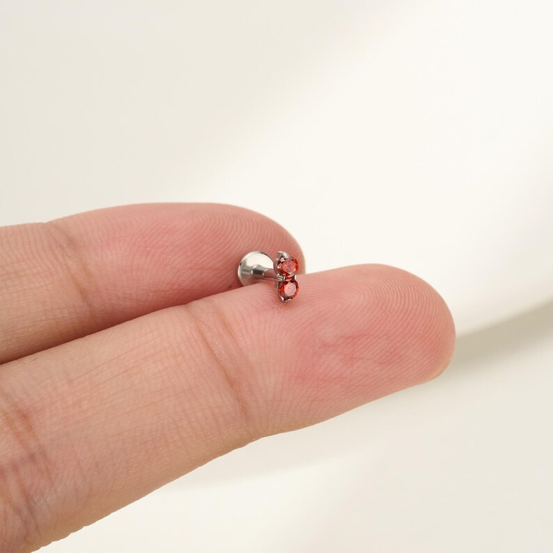 Pu&ograve; includere: Un gioiello per il corpo in metallo color argento con due piccole pietre preziose rotonde di colore rosso vivace, incastonate verticalmente in una montatura a griffe, fissate a un perno con base circolare piatta, per piercing con finitura lucida.