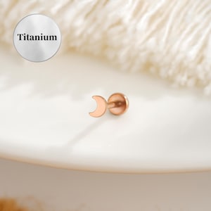 Puede incluir: Un piercing labial de oro rosa con un diseño de media luna. El perno es de titanio. La palabra "Titanium" está impresa en un círculo plateado en la esquina superior izquierda.