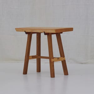 Puede incluir: Un pequeño taburete de madera hecho a mano con un asiento rectangular y cuatro patas robustas. El taburete está hecho de madera de color claro, mostrando la veta natural. Las patas están unidas por un soporte horizontal.