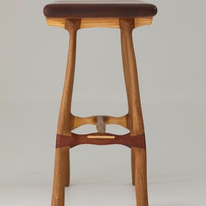 May include: A handcrafted wooden stool with a dark brown seat and light brown legs. The legs are connected by a decorative band of reddish-brown wood. The stool's design features a minimalist aesthetic, showcasing the natural wood grain.