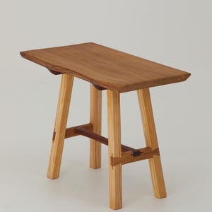 May include: A wooden stool with a rectangular seat and four angled legs. The stool is made from light-coloured wood with darker wood accents and joinery. The seat is a warm brown colour.
