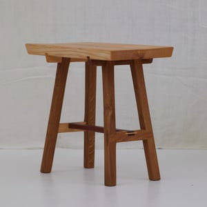 May include: A wooden stool with a rectangular seat and four angled legs. The stool is made of light-coloured wood with darker wood accents and joinery. The design is minimalist and modern, suitable for various interior styles.