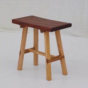 May include: A small, handcrafted wooden stool. It has a rectangular, reddish-brown seat and four light-coloured legs. The legs are joined with horizontal supports. The stool is made of different types of wood and has a natural finish.