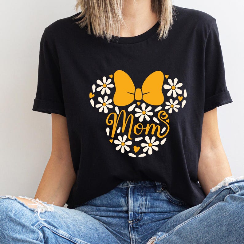 May include: Black t-shirt with a yellow bow and white flowers, forming a Mickey Mouse silhouette. The text "MOMS" is written in a stylized font inside the silhouette.