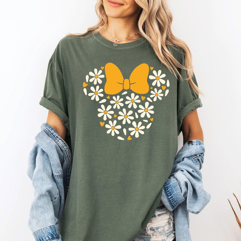May include: A green t-shirt with a white daisy and yellow bow design. The design resembles a cartoon character's head.