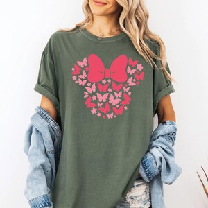 May include: A green t-shirt with a pink and red butterfly design in the shape of Mickey Mouse ears. The design features a large bow in the center.