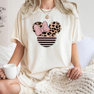 May include: A white t-shirt with a pink and black Minnie Mouse design featuring a bow, leopard print ears, and black and white stripes.