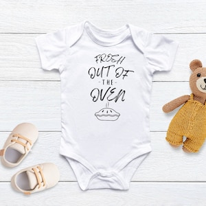 Op de afbeelding: Een witte baby romper met de tekst "Fresh Out of the Oven" en een zwart-witte illustratie van een taart. Een gehaakte bruine teddybeer bevindt zich aan de rechterkant van de afbeelding.