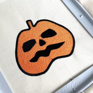 May include: A white embroidery hoop with a 120x120mm marking. Inside the hoop is a piece of white fabric with an orange and black embroidered pumpkin design.