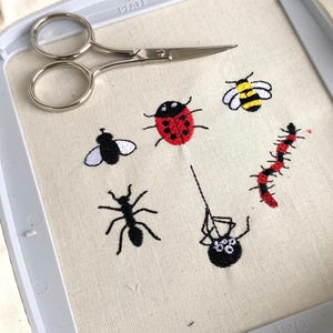Könnte beinhalten: Stickereien von einem Marienkäfer, einer Biene, einer Fliege, einer Ameise, einer Spinne und einer rot-schwarzen Raupe auf einem weißen Stoff.