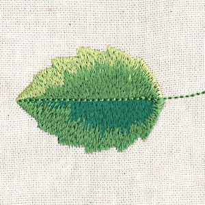 May include: A green embroidered leaf with a light green outline on a white fabric background.