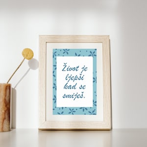 Könnte beinhalten: Ein gerahmter Druck mit dem Text "Život je ljepši kad se smiješ" in blauer Schrift. Der Druck hat einen hellblauen floralen Rand und ist in einem hellen Holzrahmen gefasst. Eine getrocknete Blume und eine Vase sind im Hintergrund.