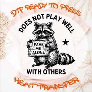 May include: A light pink t-shirt featuring a black graphic of a raccoon holding a wooden sign that reads LEAVE ME ALONE. The text DOES NOT PLAY WELL WITH OTHERS arches around the raccoon, accompanied by two black stars on either side of the animal.