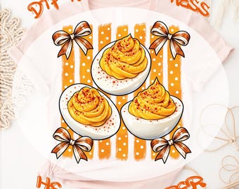 Op de afbeelding: Een lichtroze t-shirt met een afbeelding van drie gevulde eieren met oranje paprikapoeder, omringd door vier oranje-witte gestreepte strikken. De tekst DTF READY TO PRESS HEAT TRANSFER staat in het oranje boven en onder het ontwerp.