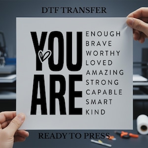 May include: A white DTF transfer sheet with the words "YOU ARE" in large black letters, a heart graphic, and a list of positive affirmations. The text "DTF TRANSFER" and "READY TO PRESS" are also printed on the sheet.