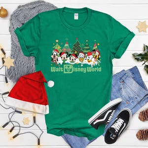 Green Christmas t-shirt featuring Mickey Mouse and friends in front of Christmas trees. The text on the shirt says 'Walt Disney World'.  The shirt is shown on a flat surface with a Santa hat, jeans, and Vans shoes.