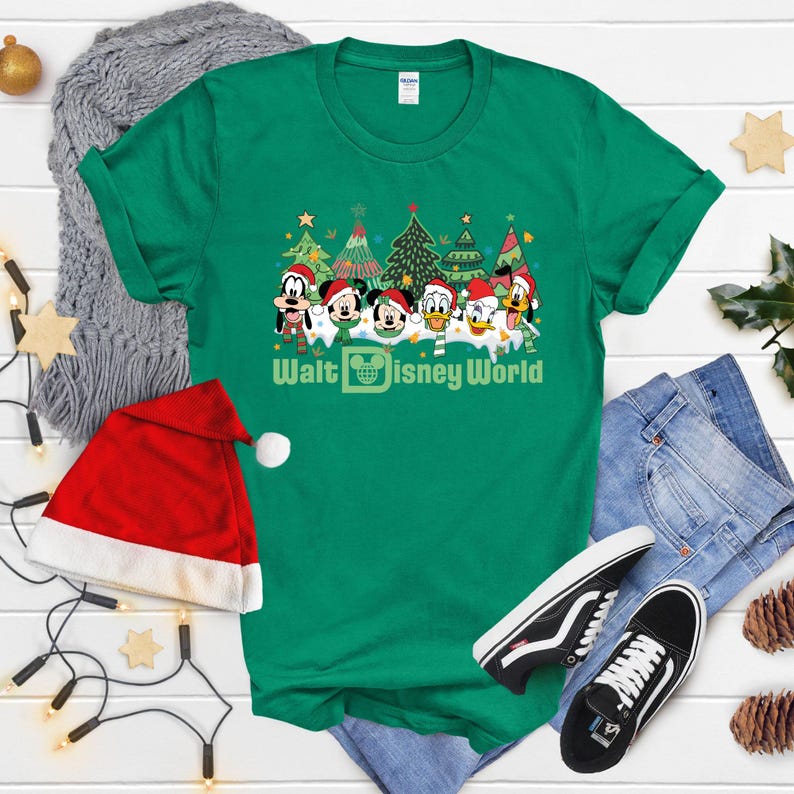 Green Christmas t-shirt featuring Mickey Mouse and friends in front of Christmas trees. The text on the shirt says 'Walt Disney World'.  The shirt is shown on a flat surface with a Santa hat, jeans, and Vans shoes.