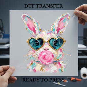 May include: DTF transfer sheet featuring a colorful bunny with heart-shaped sunglasses and a pink bubblegum bubble. The bunny is painted with textured brushstrokes in shades of pink, blue, and gold. Text at the top reads "DTF TRANSFER" and at the bottom "READY TO PRESS".