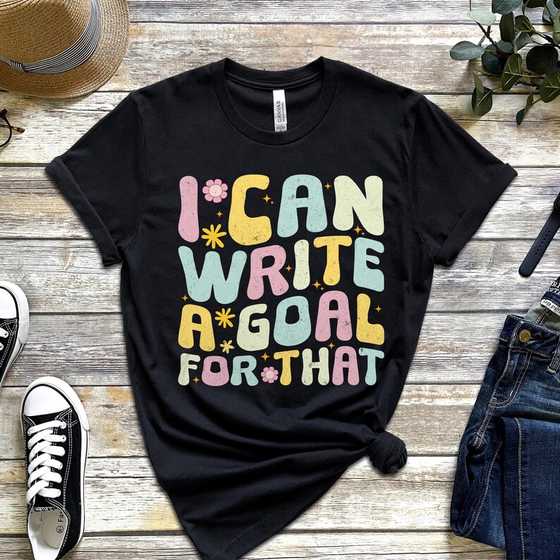 Black t-shirt with the text "I CAN WRITE A GOAL FOR THAT" in colorful, retro-style lettering. The design includes flowers and stars. The shirt is made of a soft material.