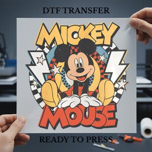 May include: A DTF transfer sheet featuring a smiling Mickey Mouse graphic. The design includes the words "Mickey Mouse" in bold, colorful lettering, with lightning bolt and star accents. The text "Ready to Press" is also visible.