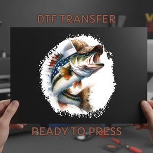 May include: A black DTF transfer sheet featuring a leaping bass wearing a trucker hat with an American flag design. The fish is detailed with realistic scales and fins, set against a white splash. Text at the top reads "DTF TRANSFER" and "READY TO PRESS" at the bottom.
