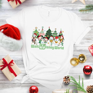 White Christmas t-shirt featuring Mickey Mouse and friends in front of Christmas trees. The shirt has green text that says 'Walt Disney World'.  The characters include Mickey, Minnie, Goofy, Donald, and Pluto.  The design is festive and features cartoon characters in a winter setting.