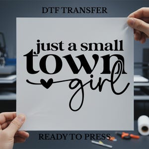 May include: A white DTF transfer sheet with the text "just a small town girl" in black script. The words are arranged in a stacked format with a heart symbol. The words "DTF TRANSFER" and "READY TO PRESS" are also printed on the sheet.