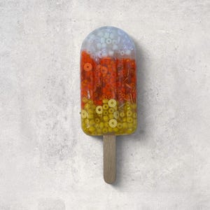 May include: A colorful popsicle-shaped art piece with a wooden stick. The top section is filled with white and light blue beads, transitioning to orange and yellow beads. The artwork is set against a light gray background.