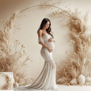 May include: A pregnant person wearing a white lace gown stands in front of a backdrop of dried pampas grass and a gold hoop. The person is holding their belly and looking down.